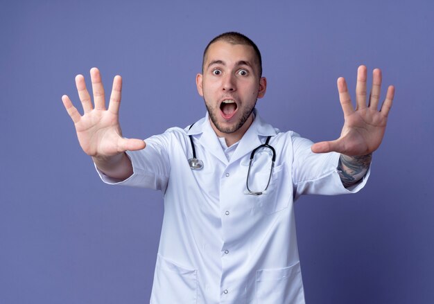 Impressed young male doctor wearing medical robe and stethoscope around his neck stretching out hands gesturing stop isolated on purple