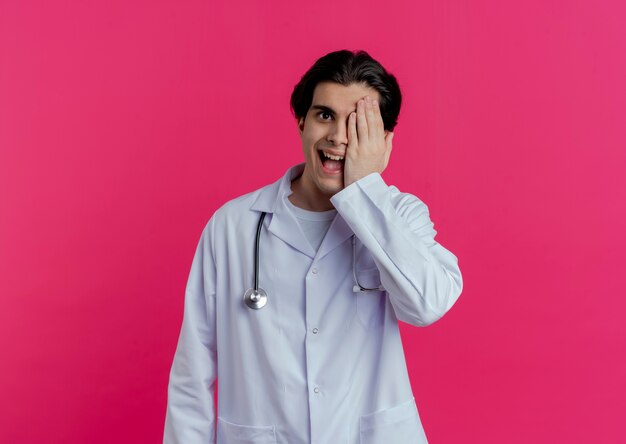 Impressed young male doctor wearing medical robe and stethoscope  covering half of face isolated on pink wall with copy space