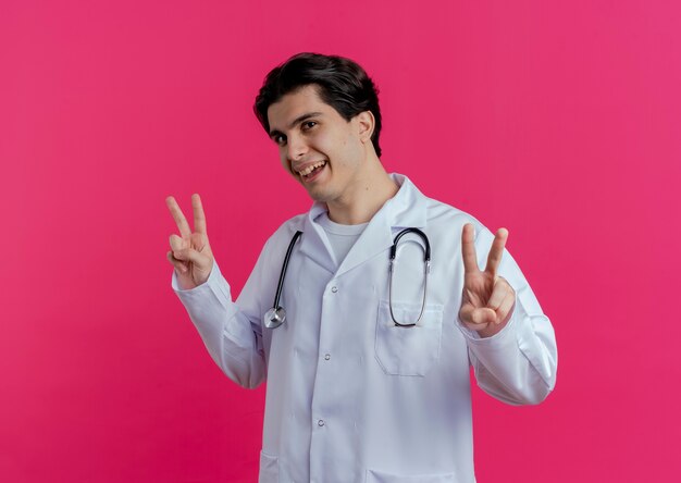 Impressed young male doctor wearing medical robe and stethoscope  doing peace sign isolated on pink wall with copy space