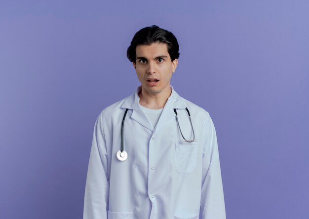 Impressed young male doctor wearing medical robe and stethoscope  isolated on purple wall with copy space