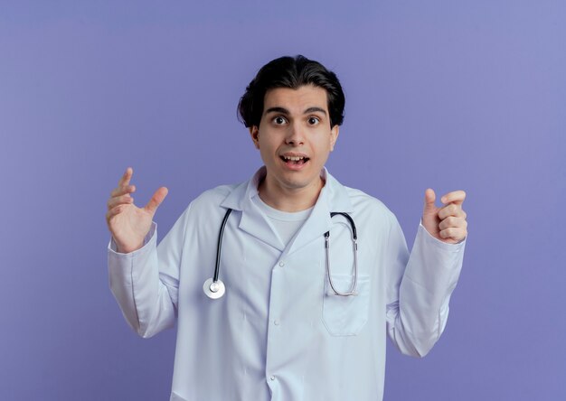 Впечатленный молодой мужчина-врач в медицинском халате и стетоскопе смотрит, держа руки в воздухе, держа руки изолированными