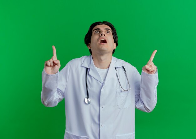 Впечатленный молодой мужчина-врач в медицинском халате и стетоскопе смотрит и указывает вверх изолированно на зеленой стене с копией пространства