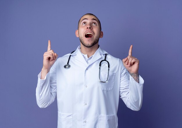 Impressed young male doctor wearing medical robe and stethoscope looking and pointing up isolated on purple