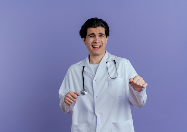 Впечатленный молодой мужчина-врач в медицинском халате и стетоскопе смотрит в сторону, держа руки в воздухе изолированными