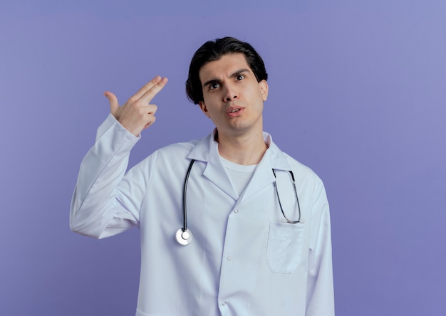 Impressed young male doctor wearing medical robe and stethoscope looking up doing pistol gesture isolated on purple wall with copy space