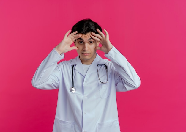 의료 가운 및 복사 공간이 분홍색 벽에 고립 된 큰 눈을 만드는 청진기를 착용하는 감동 된 젊은 남성 의사