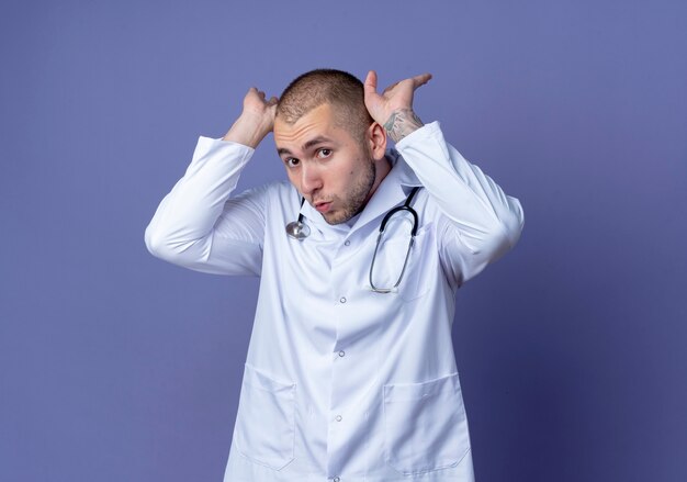 Impressed young male doctor wearing medical robe and stethoscope putting hands near head isolated on purple