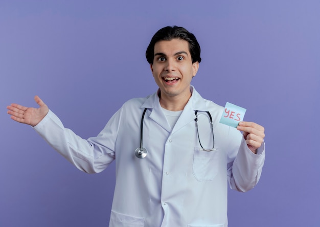 Impressed young male doctor wearing medical robe and stethoscope  showing yes note and empty hand isolated on purple wall