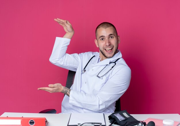 Impressed young male doctor wearing medical robe and stethoscope sitting at desk with work tools showing size with hands isolated on pink