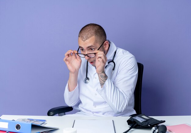 Free photo impressed young male doctor wearing medical robe and stethoscope sitting at desk with work tools wearing and holding glasses and looking at folder isolated on purple