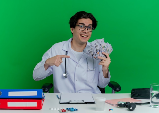 Foto gratuita impressionato giovane medico maschio che indossa veste medica e stetoscopio con gli occhiali seduto alla scrivania con strumenti medici che tengono e che punta al denaro isolato sulla parete verde