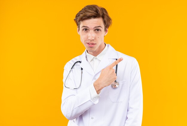 Impressed young male doctor wearing medical robe with stethoscope points at side isolated on orange wall