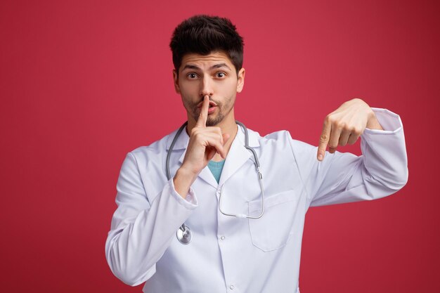 Impressed young male doctor wearing medical uniform and stethoscope around his neck looking at camera pointing down showing silence gesture isolated on red background