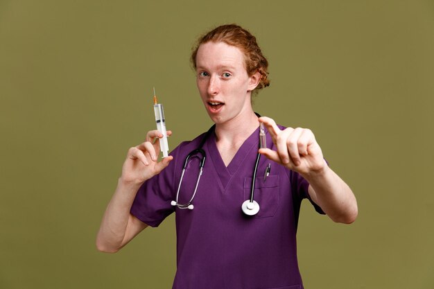 Impressed young male doctor wearing uniform with stethoscope holding syringe with ampoule isolated on green background