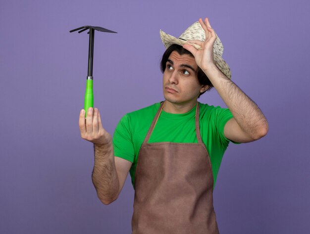 Impressed young male gardener in uniform wearing gardening hat holding and looking at hoe rake grabbed hat