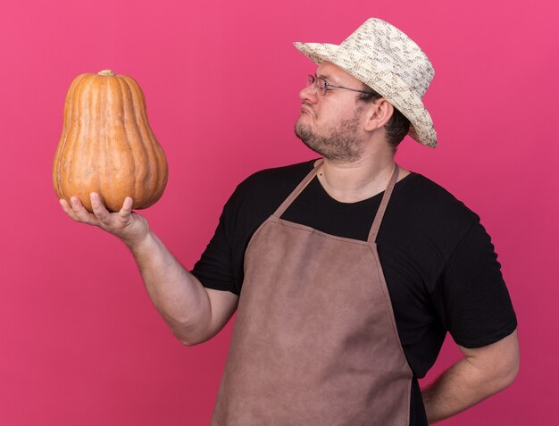 Impressionato giovane giardiniere maschio che indossa cappello da giardinaggio tenendo e guardando zucca isolato sulla parete rosa