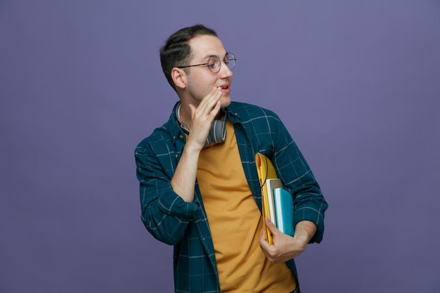impressed young male student wearing glasses headphones around neck holding folder note book note pad under arm looking at side showing whisper gesture isolated on purple background