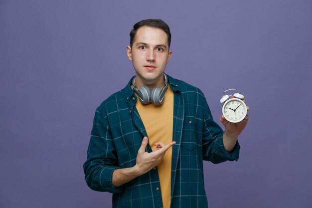 impressed young male student wearing headphones around neck looking at camera showing alarm clock pointing at it isolated on purple background
