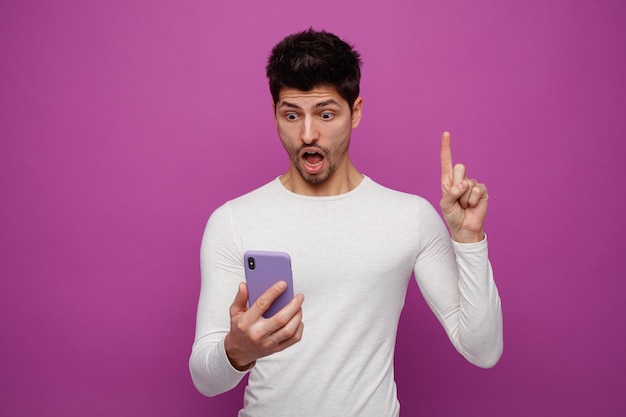 Impressed young man holding and looking at mobile phone pointing up isolated on purple background