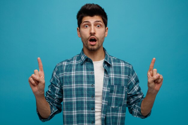 Impressed young man looking at camera pointing fingers up isolated on blue background