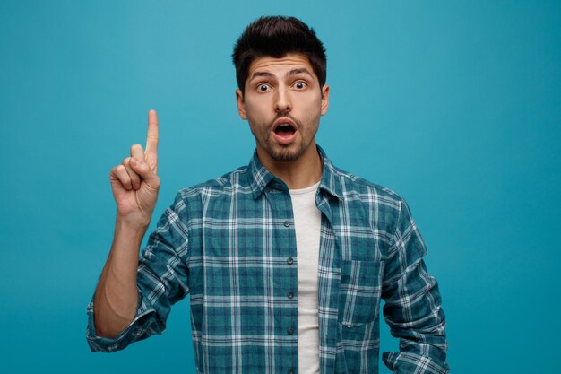 Impressed young man looking at camera pointing up isolated on blue background