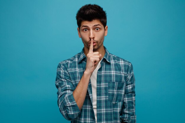 Impressed young man looking at camera showing silence gesture isolated on blue background