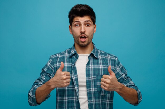 Impressed young man looking at camera showing thumbs up isolated on blue background