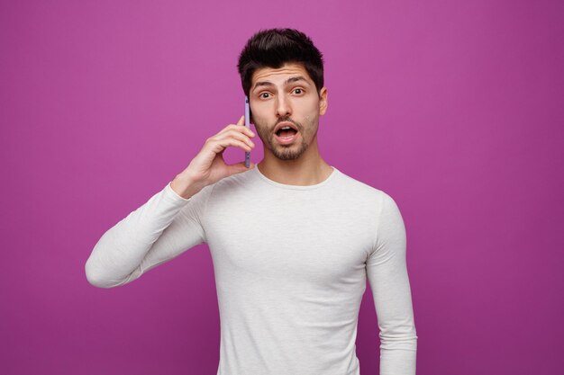 Impressed young man looking at camera talking on phone isolated on purple background