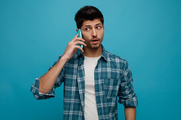 Впечатленный молодой человек разговаривает по телефону, глядя в сторону, изолированную на синем фоне