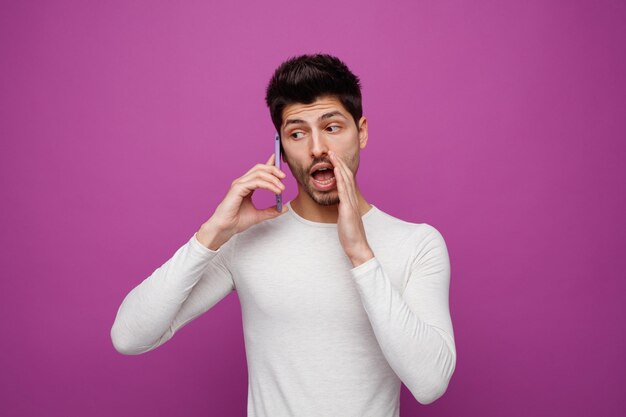 Impressed young man talking on phone looking at side keeping hand near mouth whispering isolated on purple background