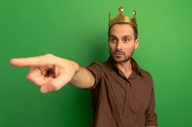 Impressed young man wearing crown looking and pointing at side isolated on green wall