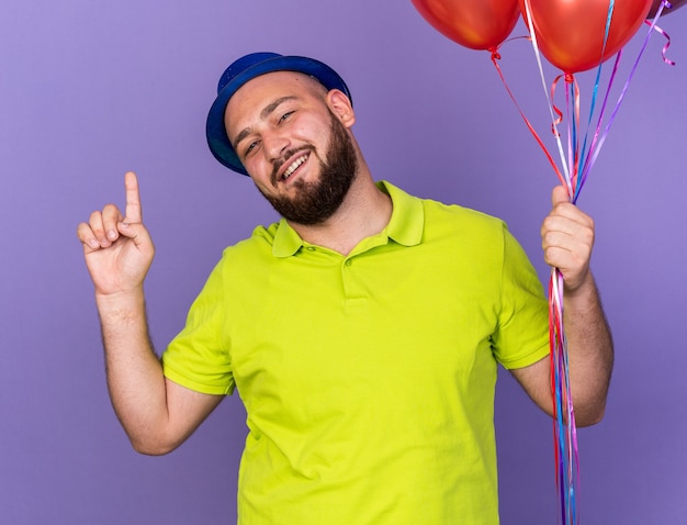 Impressed young man wearing party hat holding balloons points at up isolated on blue wall