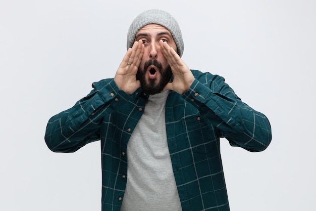 Giovane impressionato che indossa un cappello invernale guardando la fotocamera tenendo le mani vicino alla bocca chiamando qualcuno fuori isolato su sfondo bianco