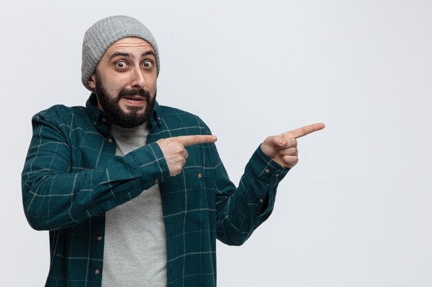 Impressed young man wearing winter hat looking at camera pointing to side isolated on white background