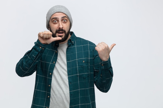 Foto gratuita giovane uomo impressionato che indossa il cappello invernale guardando la fotocamera che punta al lato isolato su sfondo bianco