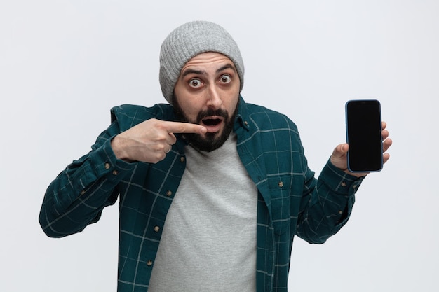 Впечатленный молодой человек в зимней шапке смотрит в камеру, показывая мобильный телефон на камеру, указывая на него на белом фоне