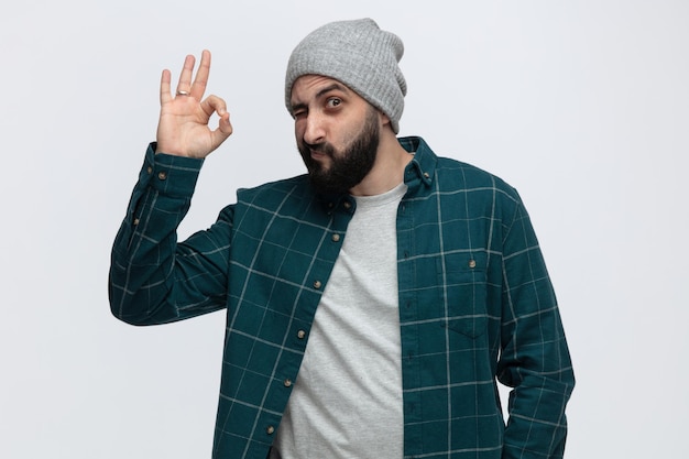 Impressed young man wearing winter hat looking at camera showing ok sign winking isolated on white background