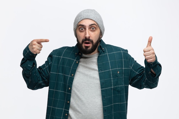 Impressed young man wearing winter hat looking at camera showing thumb up pointing at his thumb isolated on white background