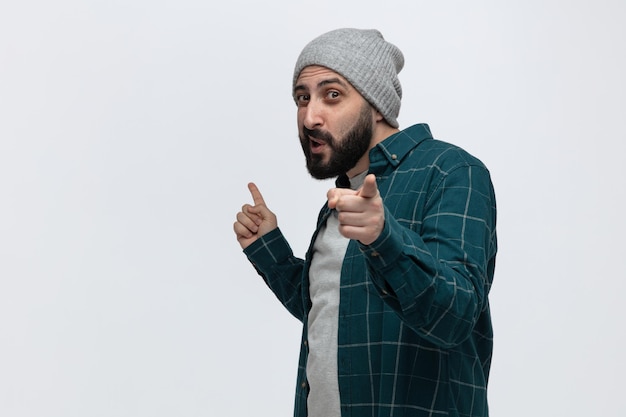 Impressed young man wearing winter hat standing in profile view looking at camera pointing up and at camera isolated on white background with copy space