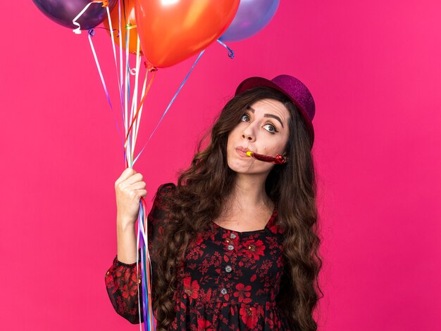 Впечатленная молодая тусовщица в партийной шляпе держит воздушные шары, дует партийный рог, глядя в сторону, изолированную на розовой стене с копией пространства