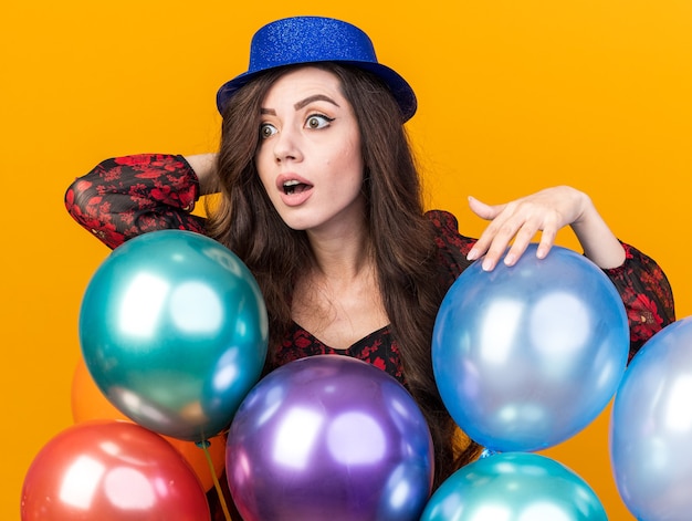 Impressionato giovane ragazza che indossa un cappello da festa in piedi dietro i palloncini che si toccano tenendo la mano dietro la testa guardando il lato isolato sul muro arancione