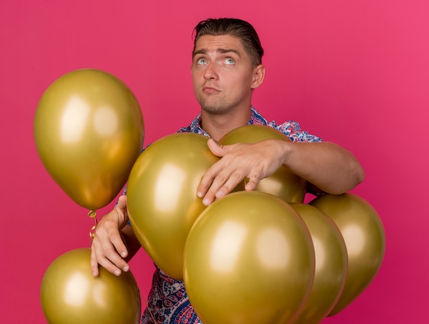 Impressed young party guy looking up wearing colorful shirt standing among balloons isolated on pink