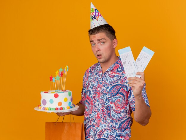 Впечатленный молодой тусовщик в кепке дня рождения держит подарочный пакет с тортом и билетами, изолированными на оранжевом