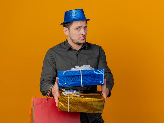 무료 사진 오렌지에 고립 된 상자와 선물 가방을 들고 파란색 모자를 쓰고 감동 된 젊은 파티 남자