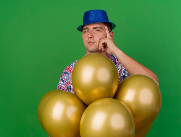 Impressed young party guy wearing blue hat standing behind balloons and putting hand on temple isolated on green