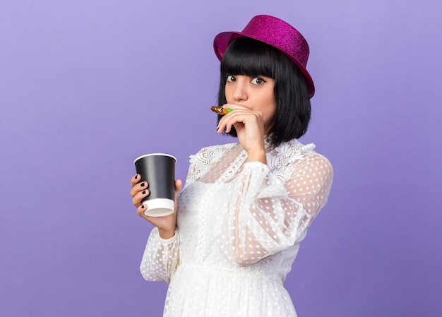 紫色の壁で隔離された正面を見て、口の中にパーティーホーンと別の手でプラスチック製のコーヒーカップを保持している縦断ビューで立っているパーティーハットを身に着けている感動の若いパーティー女性