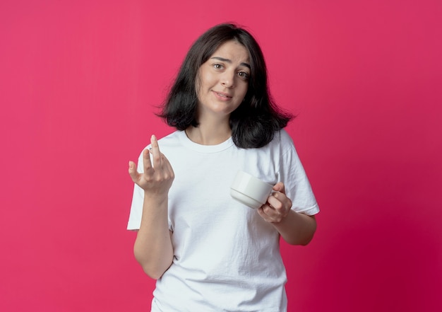 Impressionato giovane ragazza piuttosto caucasica tenendo la tazza e tenendo la mano in aria isolato su sfondo cremisi con spazio di copia