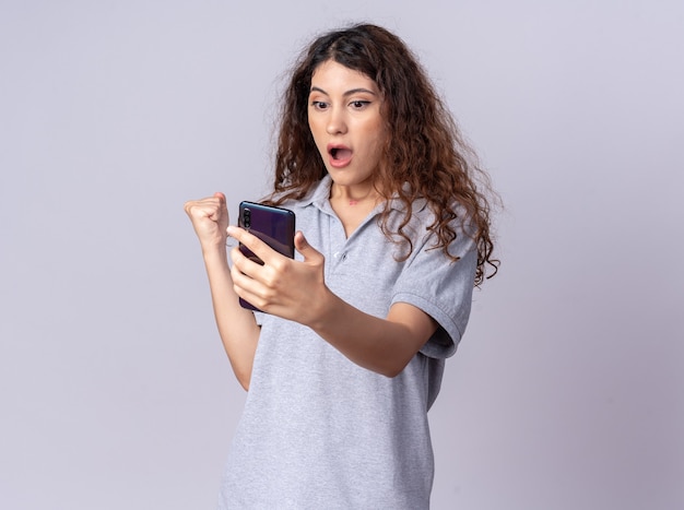 Impressed young pretty caucasian girl holding and looking at mobile phone clenching fist 