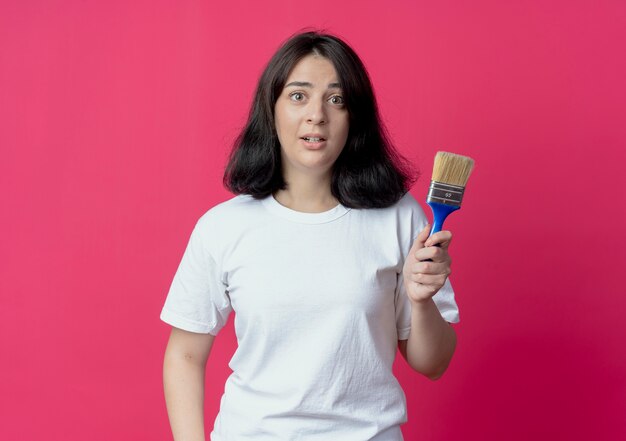 Impressed young pretty caucasian girl holding paint brush isolated on crimson background with copy space