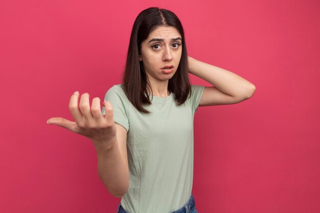 Impressed young pretty caucasian girl  keeping hand behind head stretching out another hand  isolated on pink wall